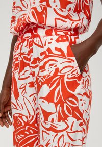 Czerwona i biała tkanina w wzór z liściami. Elastyczny pas z widoczną boczną kieszenią. Gładka, lekka tekstura.