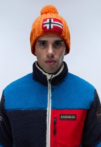Berretto lavorato a maglia arancione con pompon e design della bandiera norvegese. Il modello indossa una giacca in pile multicolore con sezioni blu, blu scuro e rosse.
