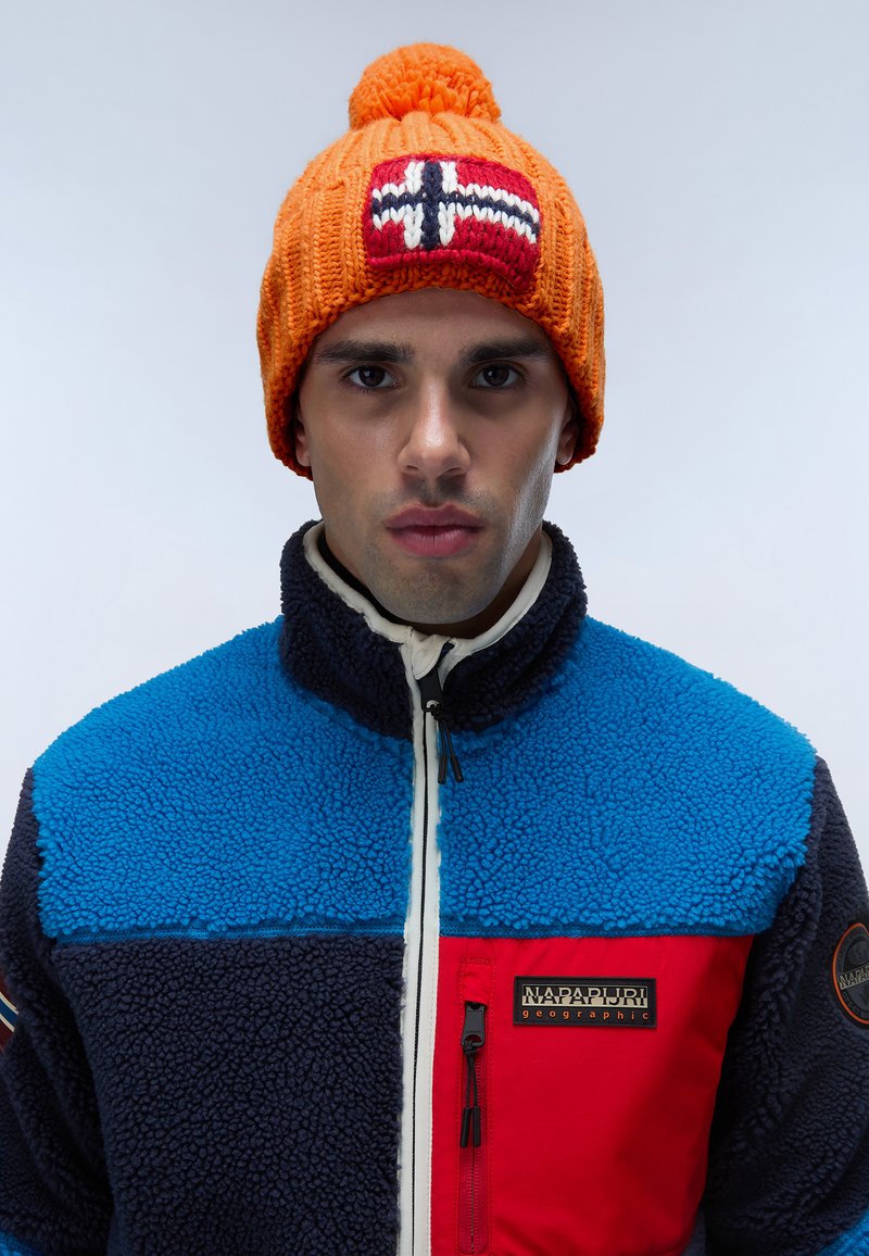 Berretto lavorato a maglia arancione con pompon e design della bandiera norvegese. Il modello indossa una giacca in pile multicolore con sezioni blu, blu scuro e rosse.