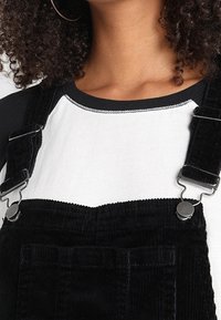 Svarta kordflöjlsbyxor med silverdetaljer, som har justerbara axelband, en framficka och en kontrasterande vit skjorta med svart krage.