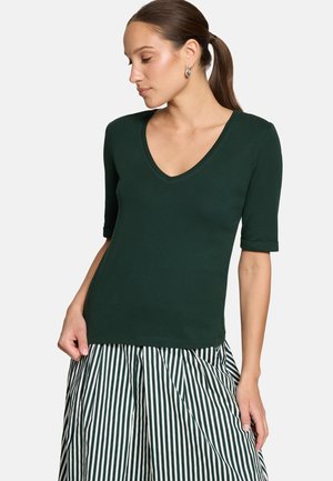 Femme avec queue de cheval brune portant un haut vert foncé à col en V et manches aux coudes, ainsi qu'une jupe à rayures verticales vertes et blanches, regardant vers la gauche.