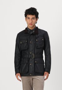 Chaqueta impermeable negra con cuello de pana marrón, cuatro bolsillos frontales, cintura ajustada con cinturón y detalles en metal dorado. Textura suave, corte entallado.