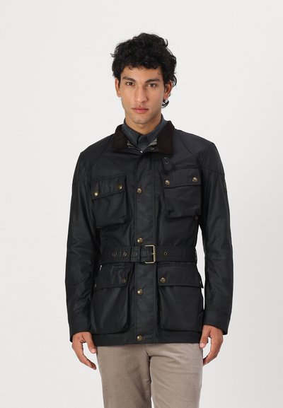 Veste imperméable noire avec col en velours côtelé marron, quatre poches avant, taille ceinturée et accents en laiton. Texture lisse, coupe ajustée.