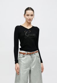 Suéter negro de manga larga con cuello en V y logo metálico "Guess" en el frente, combinado con pantalones anchos metálicos plateados y un cinturón marrón.