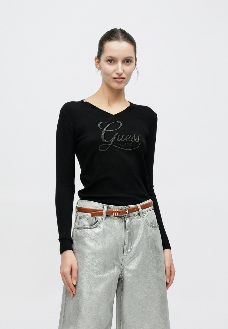 Suéter negro de manga larga con cuello en V y logo metálico "Guess" en el frente, combinado con pantalones anchos metálicos plateados y un cinturón marrón.