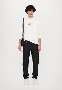 Hvid sweatshirt med sort logo, sorte jeans og hvide sneakers. Outfittet har en skuldertaske og en afslappet pasform.