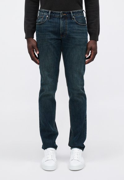 Sötétkék farmernadrágot, fehér sportcipőt és fekete hosszú ujjú pólót viselő férfi, egy sima fehér háttér előtt áll.