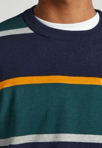 Närbild av en person som bär en marinblå, grön och grå randig tröja med ett horisontellt streck i senapsgul färg över bröstet.