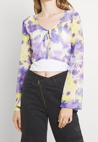 Long-sleeve, cropped top in yellow and purple tie-dye pattern with a v-neck and drawstring detail. Paired with dark pants featuring a zip.
