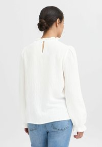 Witte, gevarieerde blouse met een ruchehalslijn en een sleutelgatdetail op de rug. Lange, opgeblazen mouwen met elastische manchetten. Combinatie met blauwe spijkerbroek.