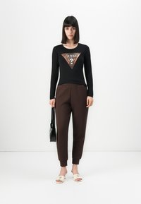 Donna con capelli lunghi e lisci di colore scuro, che indossa una maglietta nera a maniche lunghe con il logo Guess, pantaloni marroni, tacchi bianchi aperti e tiene una borsa nera.