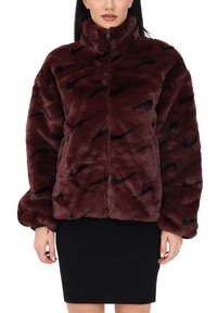 Kvinna iklädd en mörkröd fuskpälsjacka med svarta Nike-swoosh-mönster och en svart kjol, stående mot en vit bakgrund.