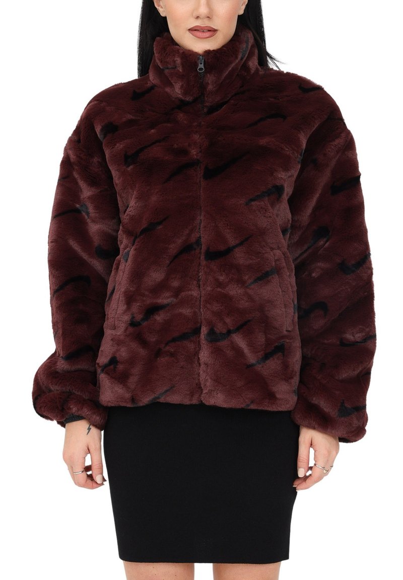 Femme portant une veste en fausse fourrure rouge foncé avec des motifs de swoosh Nike noirs et une jupe noire, se tenant devant un fond blanc.