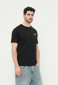 Άνδρας με σκούρα μαλλιά, που φοράει μαύρο μπλουζάκι με μικρό λευκό λογότυπο στο στήθος και ανοιχτόχρωμο μπλε τζιν, στέκεται μπροστά σε λευκό φόντο.