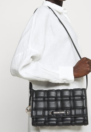 Henkilö, joka pitää kädessään mustaa, kudottua nahkaista Michael Korsin olkalaukkua, jossa on kultainen logokoru, ja jolla on valkoinen pitkähihainen paita.