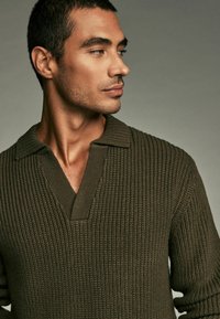 Olivovo zelený pletený sveter s výstrihom do V a rebrovanou textúrou. Má golierový výstrih a celoplošný textúrovaný vzor.