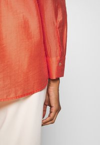 Camicia a righe rosse con una texture liscia, polsini abbottonati, orlo arrotondato e maniche lunghe, abbinata a un tessuto di colore chiaro sottostante.
