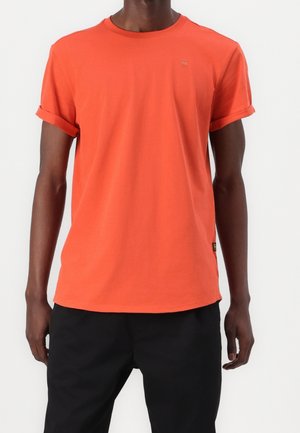 Mand iført en ensfarvet orange t-shirt med korte ærmer og rullede manchetter samt sorte bukser, stående foran en hvid baggrund.