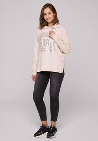 Felpa con cappuccio di colore rosa chiaro con una grafica testurizzata che recita "SPAW DAY". Abbinata a leggings grigi scuri e sneakers nere.