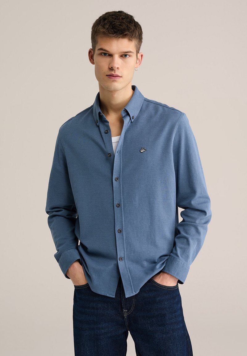 Camisa azul de manga larga con botones y tela texturizada. Presenta un pequeño logotipo bordado en el pecho y botones oscuros. Se lleva con jeans oscuros.