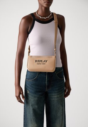Beige lærveske med kryssbody-design, preget "REPLAY"-logo, justerbar stropper og glatt tekstur. Brukes med en tettsittende tanktop og vide jeans.