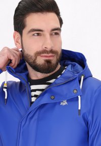 Niebieska wodoodporna kurtka z zamkiem z przodu, spiczastym kołnierzem i białymi sznurkami do ściągania. Posiada logo, napy i gładką teksturę.