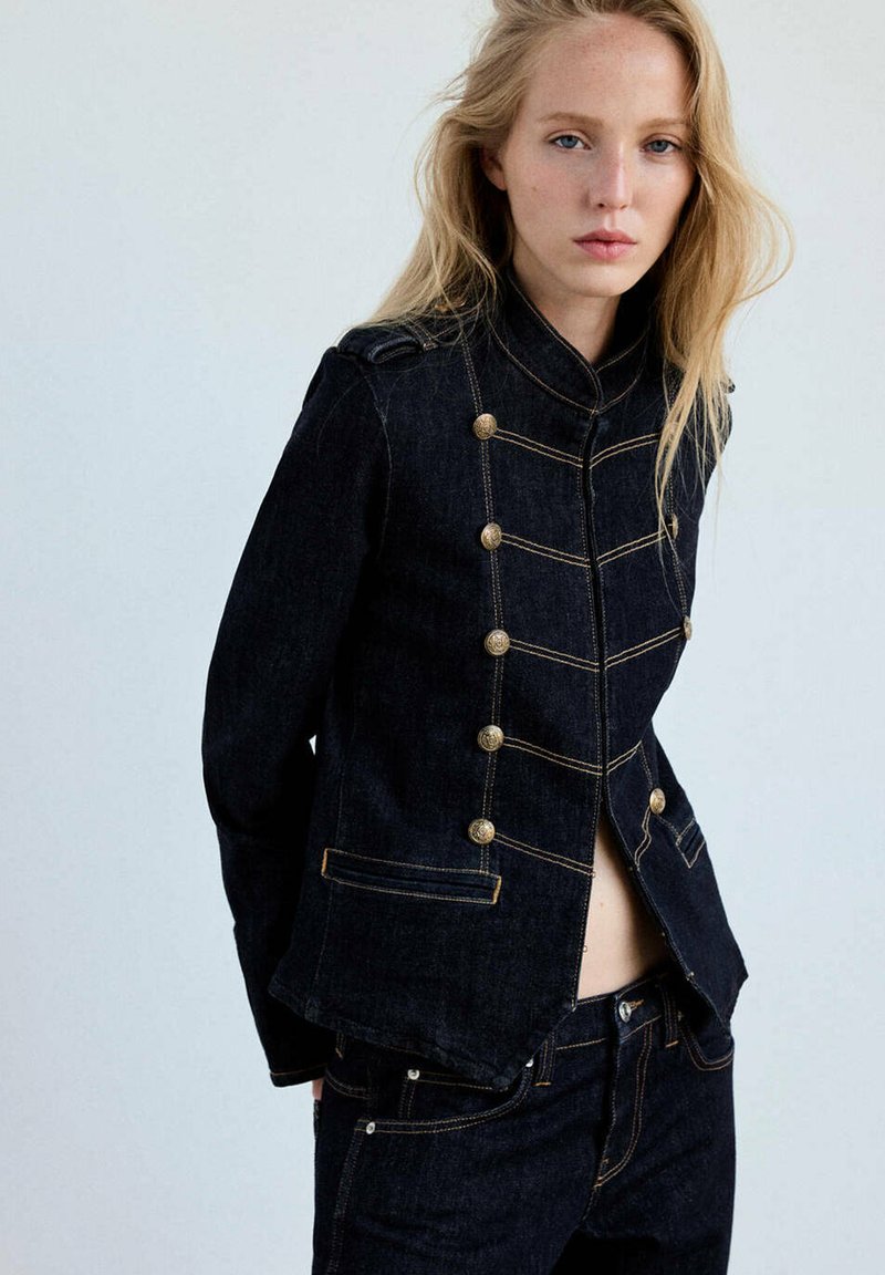 Femme blonde portant une veste militaire en denim foncé avec des boutons dorés et un jean assorti sur un fond clair uni.