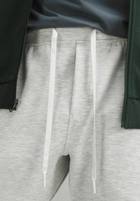 Grijze sweatpants met witte koordjes, gestructureerde stof, gecombineerd met een donkergroene ritsjack en een grijze t-shirt eronder.