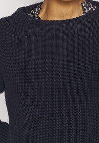 Teksturowany sweter w ciemnym granatowym kolorze z prążkowanym wzorem. Posiada koronkowy detal w kwiatowy wzór na dekolcie, który dodaje kontrastu i delikatności.