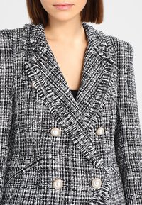 Žena v černobílém kostkovaném dvouřadém saku s texturovanými třásňovými klopami a perlami podobnými knoflíkům.