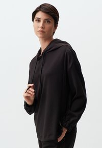 Hoodie noir en tissu doux, avec une capuche à cordon de serrage, coupe décontractée et poignets froncés. Texture lisse avec un minimum de détails.