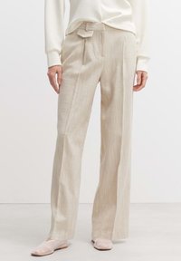 Beige, weit geschnittene Hose aus strukturiertem Stoff mit Frontfalten und einem gebundenen Bund, kombiniert mit hellrosa Slippern.
