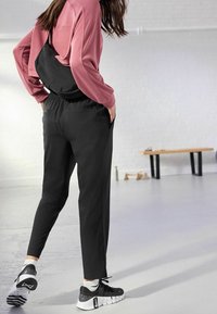 Joggers noirs fuselés avec taille élastique, associés à un haut à manches longues couleur rose et des chaussures de sport noires. Sac d'épaule noir porté.