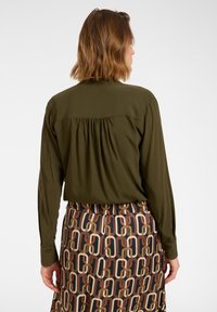 Blusa verde oliva a maniche lunghe con schiena plissettata, abbinata a una gonna con motivo a forme intrecciate in marrone, crema e blu navy.