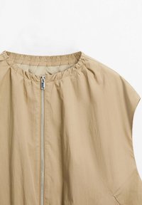 Haut beige sans manches avec encolure froncée et fermeture éclair argentée à l'avant sur fond blanc.