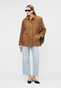 Žena v hnedom semišovom saku s strapcami, svetlomodrých širokých džínsoch, čiernych slnečných okuliaroch a čiernych špicatých lodičkách na nízkom podpätku, stojaca na bielom pozadí.