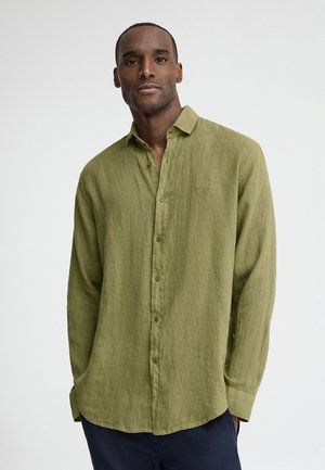 Man draagt een olijfgroen overhemd met lange mouwen en kraag en marineblauwe broek, staand tegen een effen lichte achtergrond.