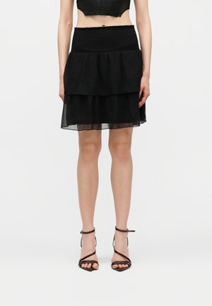 Kobieta w czarnej mini spódnicy z falbanami i marszczonym paskiem oraz czarnych, paskowanych sandałach na wysokim obcasie, stojąca na jednolitym tle.