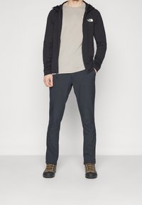 Männliches Outdoor-Outfit mit einem schwarzen Reißverschluss-Hoodie, einem hellgrauen T-Shirt, dunklen Hosen und schwarzen Wanderschuhen mit orangefarbenen Akzenten.