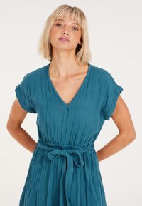 Turquoise, korte mouw jurk met een V-hals en strik taille. Gemaakt van textuurstof met verticale plooien, met een ontspannen pasvorm en een vloeiende rok.
