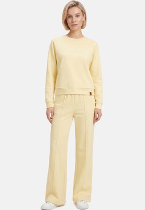 Kobieta z krótkimi blond włosami stoi, ubrana w jasnożółtą bluzę i szerokie spodnie, w połączeniu z białymi tenisówkami, na białym tle.