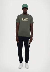 Grønn bomull T-skjorte med gull grafisk logo, kombinert med marineblå joggebukser med hvit logo-detalj. Avsluttet med hvite sneakers og en grønn caps.