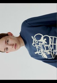 Jeune personne à la peau claire et aux cheveux bruns portant un sweat-shirt bleu marine avec un dessin abstrait blanc en lignes sur le devant.