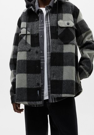 Mand iført en sort og grå ternet fleecejakke over en sort og hvid ternet skjorte og en hvid T-shirt, med sorte bukser.