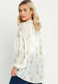 Blusa bianca trasparente con motivi floreali pastello e pois, con maniche lunghe a sbuffo e colletto arricciato, realizzata in tessuto leggero.