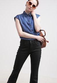 Jeansskjorte med knapper foran og oppbrettede ermer, kombinert med svarte bell-bottom bukser. Tilbehøret inkluderer en brun veske og store solbriller.