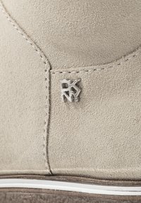 Calzado de ante beige con superficie texturizada, un pequeño logotipo plateado y costuras blancas a lo largo del borde de la suela.