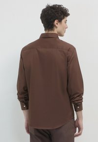 Mann mit lockigem Haar, der ein braunes langärmeliges Hemd mit Knöpfen und passende Hose trägt, von hinten vor einem einfarbigen Hintergrund gesehen.