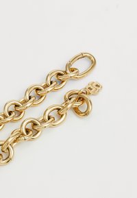 Corrente de metal dourado com grandes elos entrelaçados, apresentando uma superfície lisa e brilhante. Inclui um fecho seguro com um detalhe discreto do logótipo.