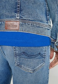 Denim jacket with brown leather label, light blue wash, and visible stitching. Blue T-shirt underneath with smooth texture. Light blue jeans with back pocket detail.